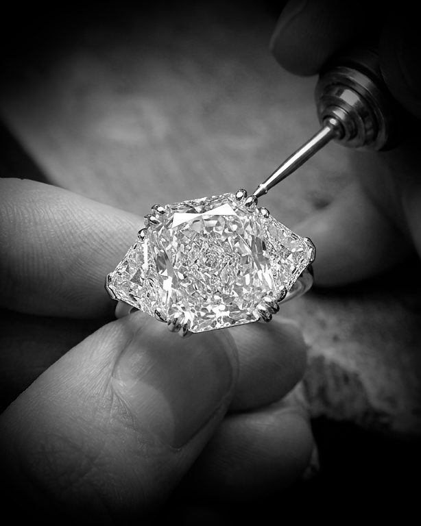Close-up of a large, square-cut diamond being prong-set into an engagement ring by a jeweler's tool.