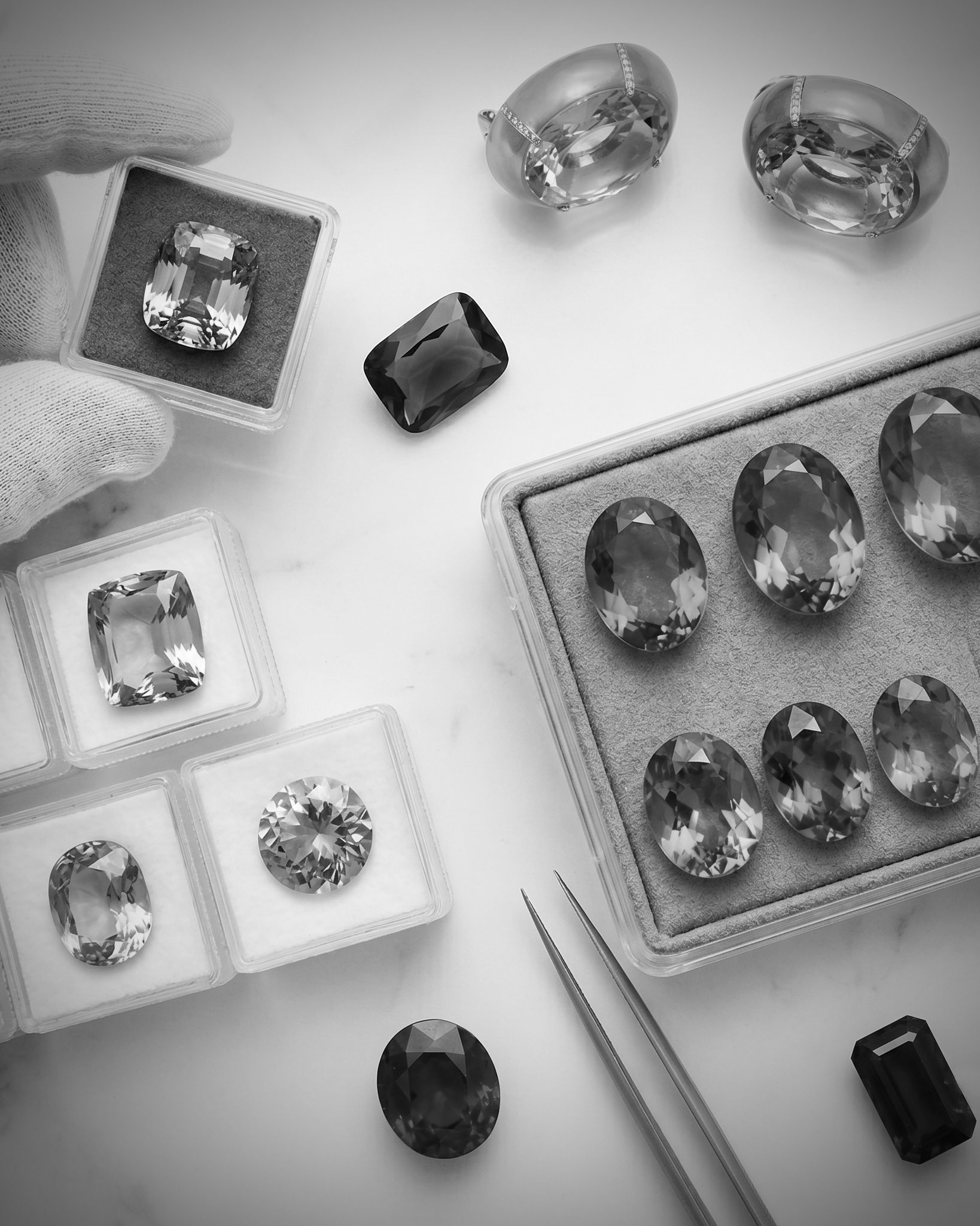 Assorted loose gemstones and jewelry pieces arranged in display boxes on a white surface (black-and-white photo).
