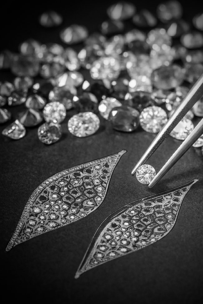 Close-up of a round diamond being picked with tweezers over two leaf-shaped, diamond-encrusted jewelry designs on a dark surface, black-and-white image.