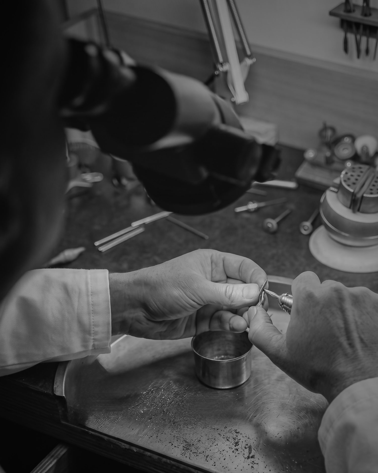 Hands used to shape a metal ring with jewelry pliers at a cluttered workbench (black and white).