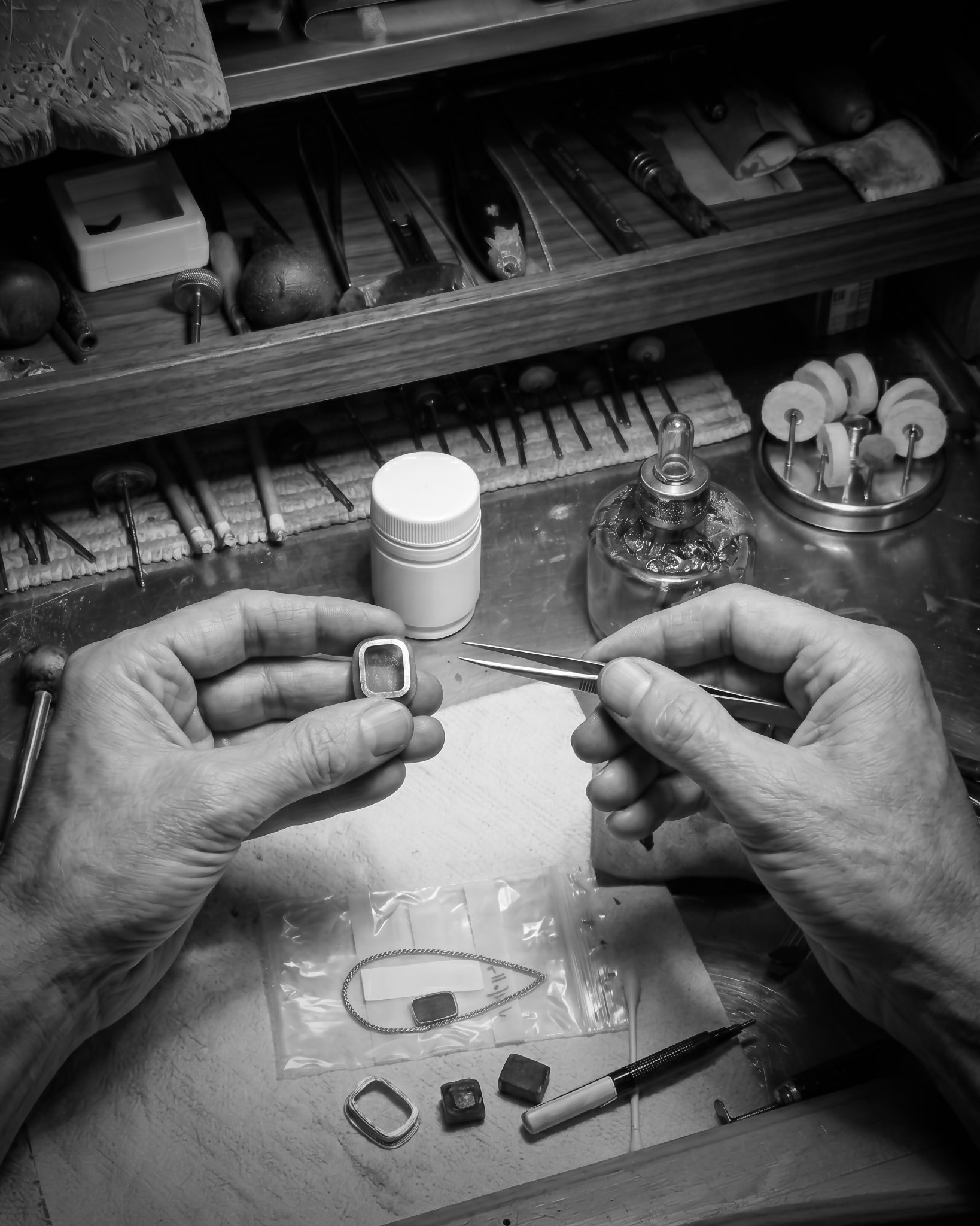 Close-up of hands assembling a small metal bezel in a jewelry workspace, with tweezers and tools on a cluttered bench.