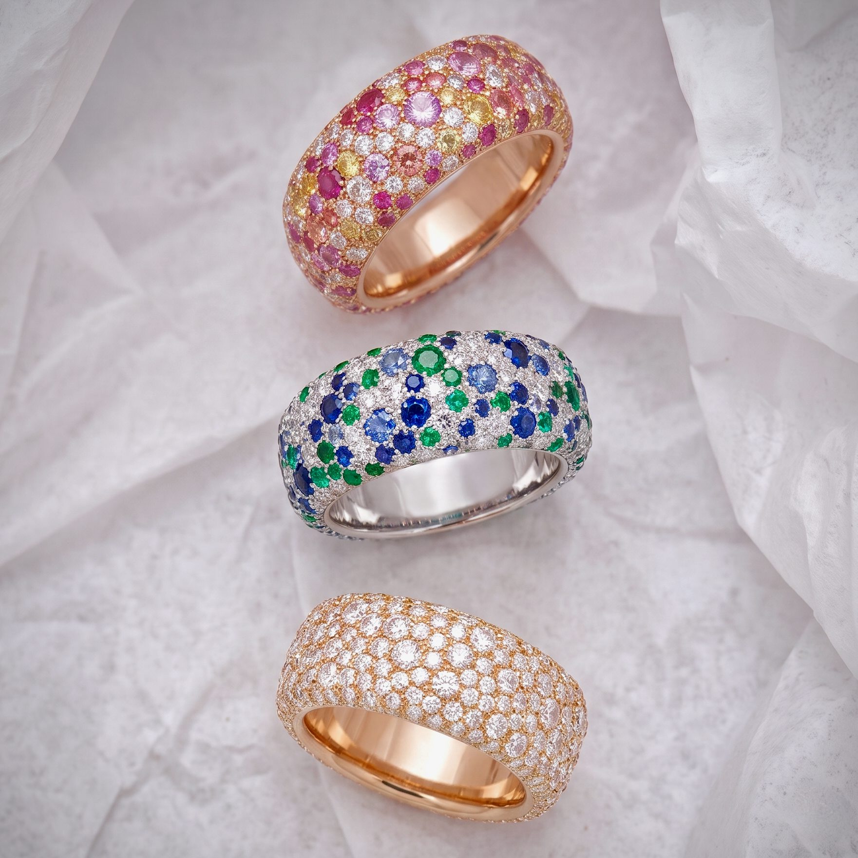 Three rose-gold rings encrusted with multicolored diamonds resting on crumpled tissue paper.