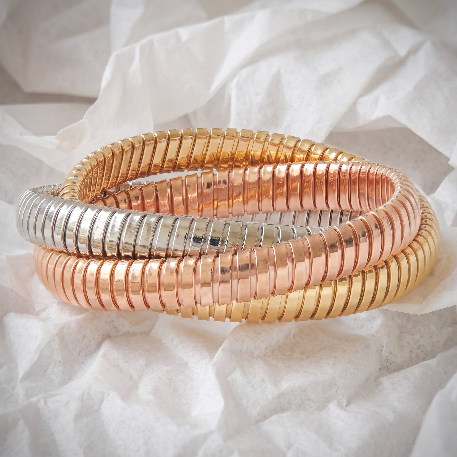 Close-up of intertwined rose gold and silver coiled bracelets resting on crumpled white tissue paper.