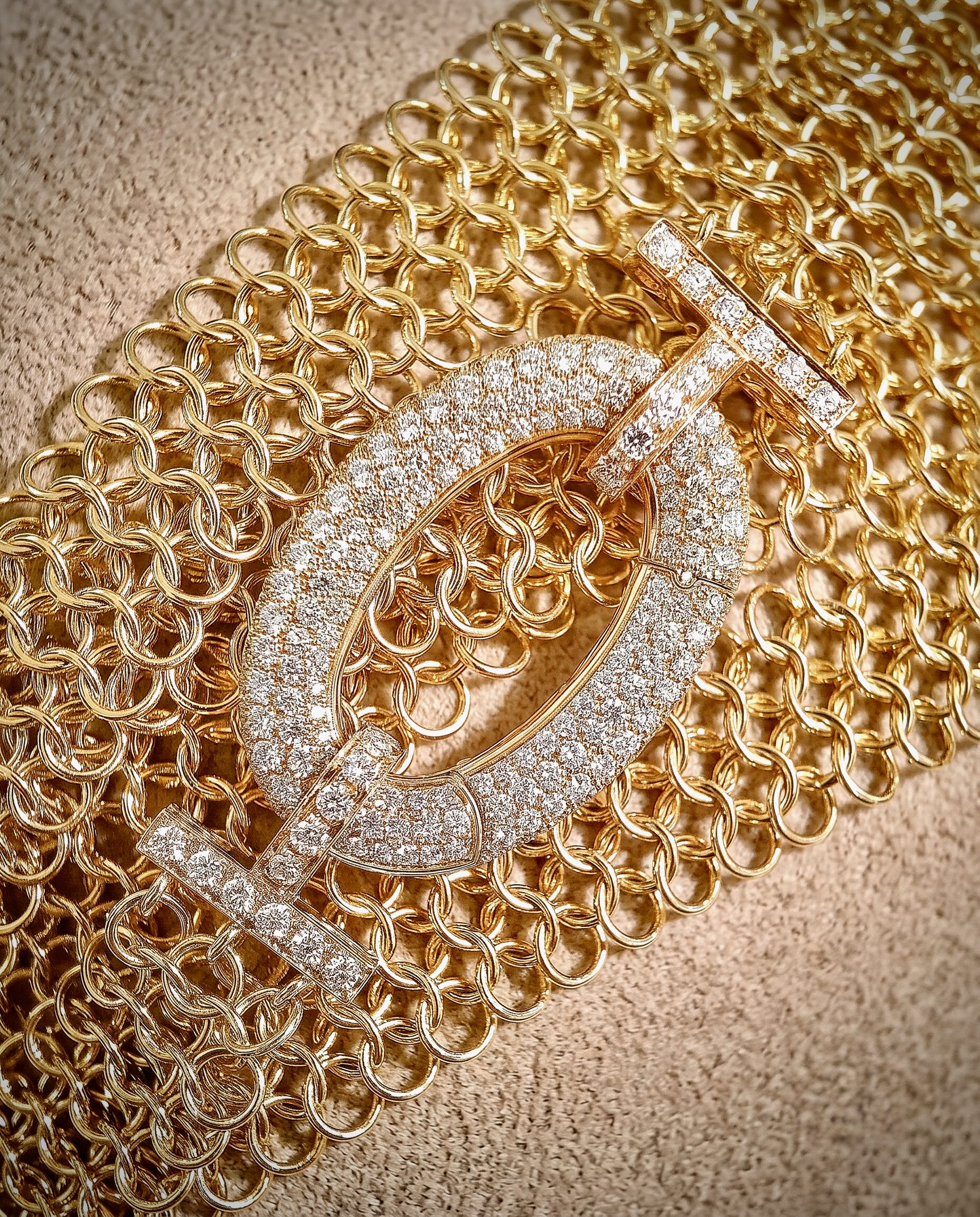 Close-up of a gold chainmail-style necklace laid on a beige carpet, showing large interlocking links and a glittery surface on some links.