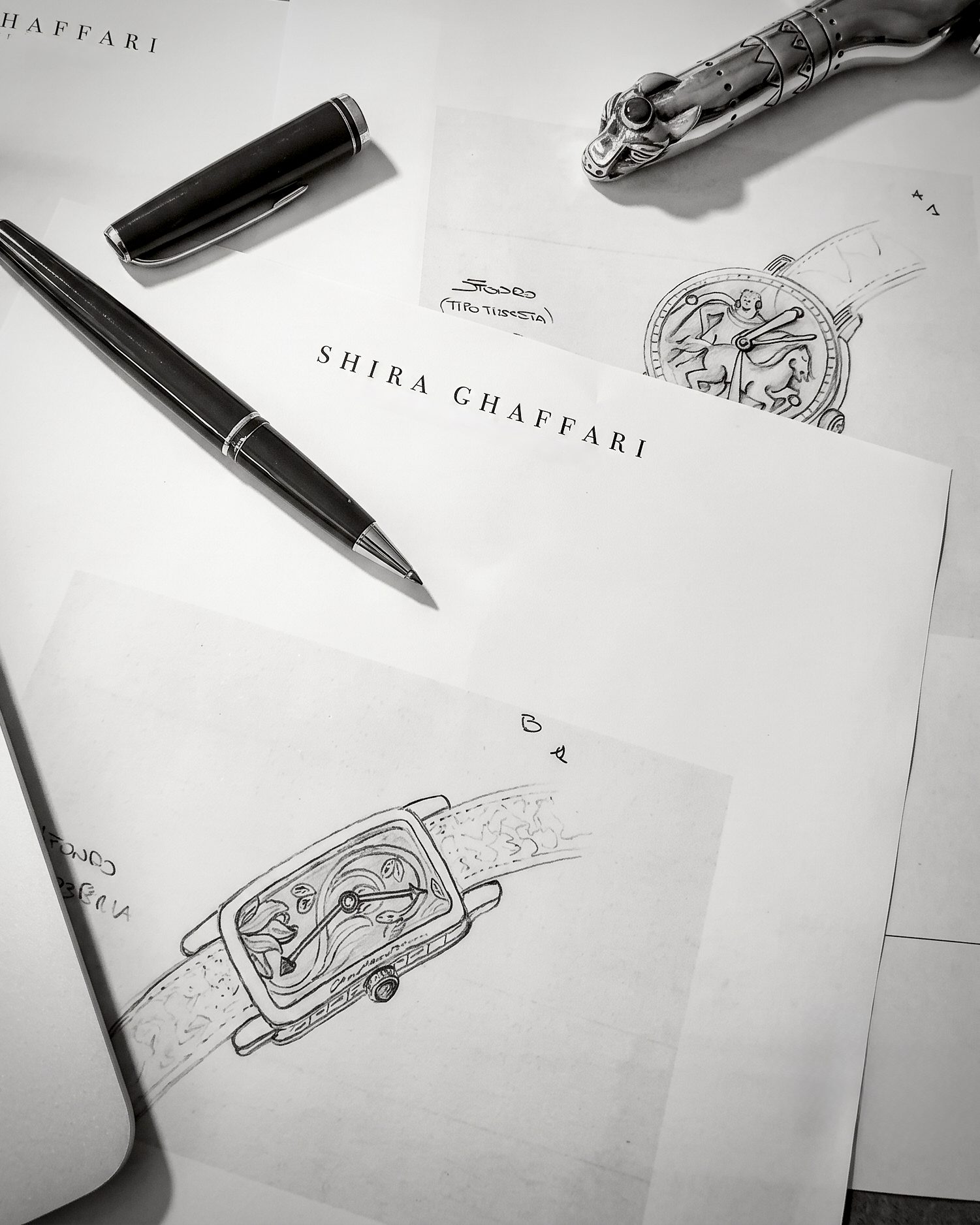 Monochrome photo of design papers labeled SHIRA GHAFFARI with two black pens and sketches of a watch and a bracelet/ornament on a table.