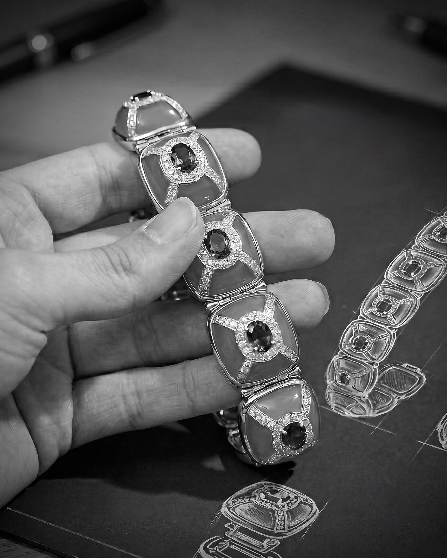 Close-up of a hand holding a silver bracelet with a dark central stone and diamond accents, with more bracelets and design sketches on a dark table nearby.