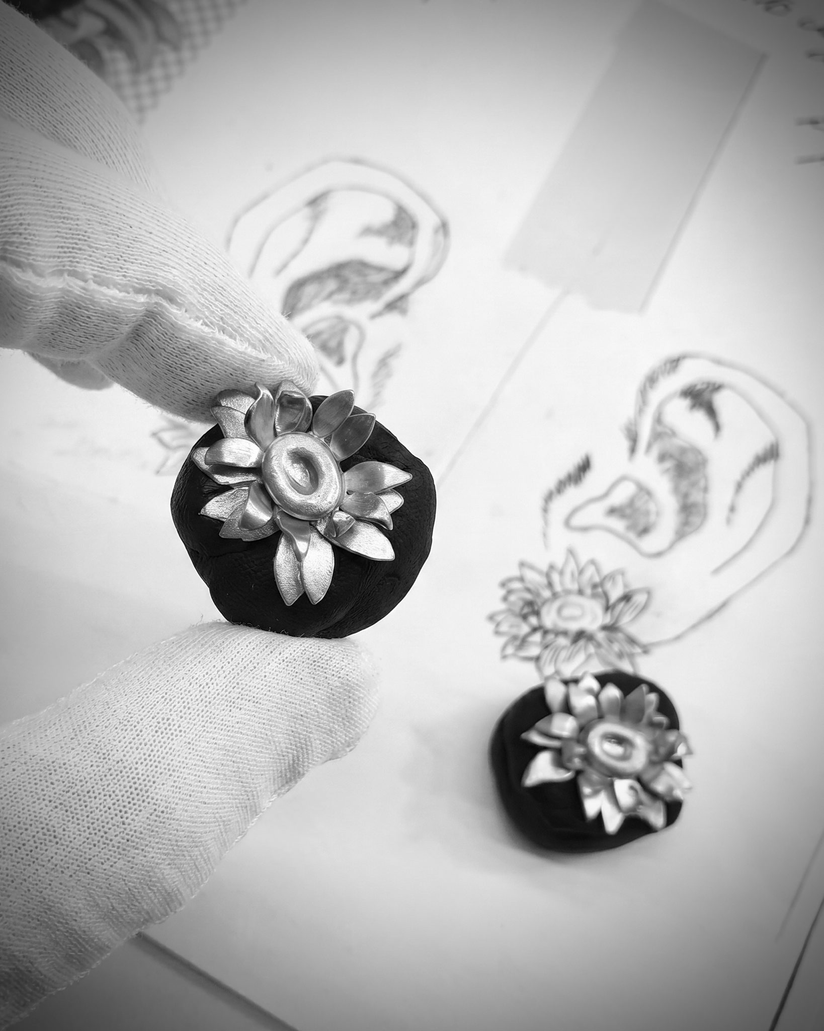 Close-up of gloved hands holding a small black velvet ring box with a metallic flower ribbon, with two other boxes in the blurred background on a sketchbook.