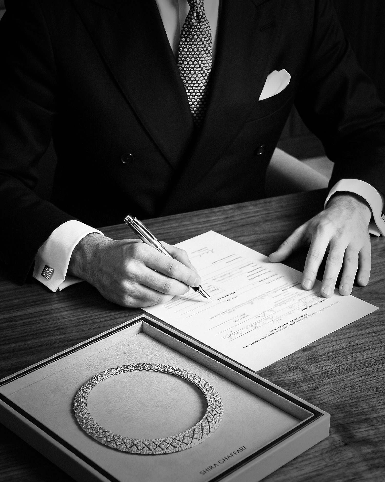 Businessperson in a suit signs a document with a pen at a desk, a decorative circular necklace sits in an open box nearby