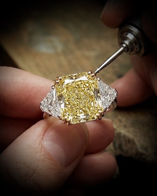 Close-up of a yellow-cut diamond ring held by fingers as a jeweler uses a rotary tool on the setting/polishing the stone.