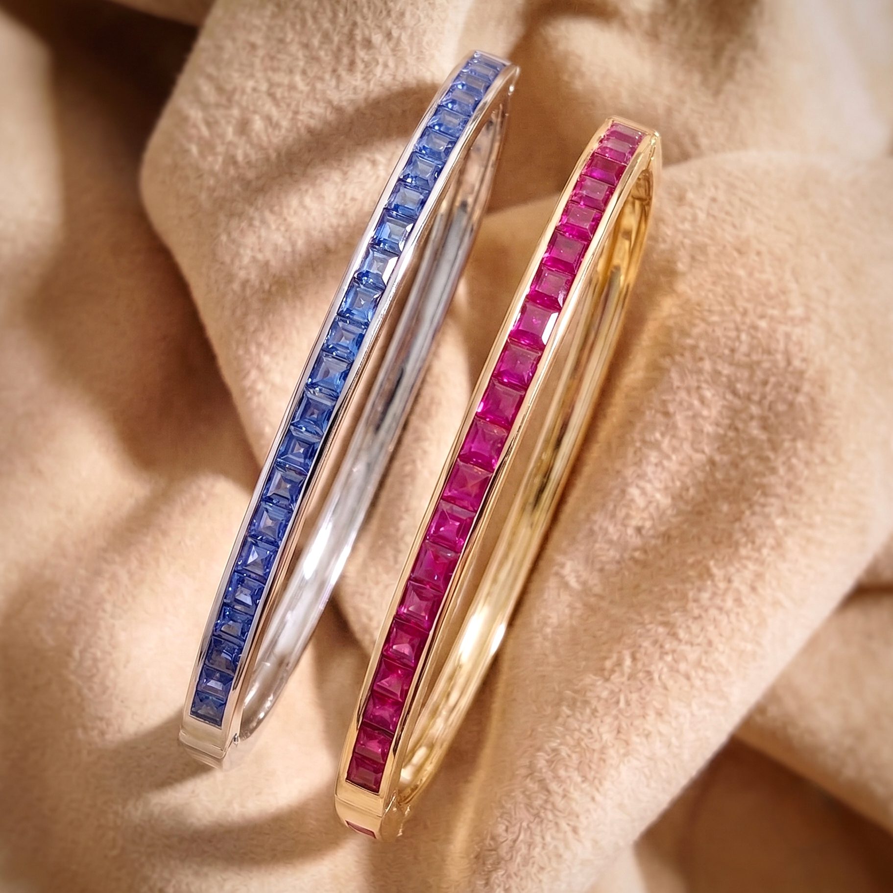 Close-up of two gold bracelets set with blue and pink square-cut gemstones on beige fabric backdrop