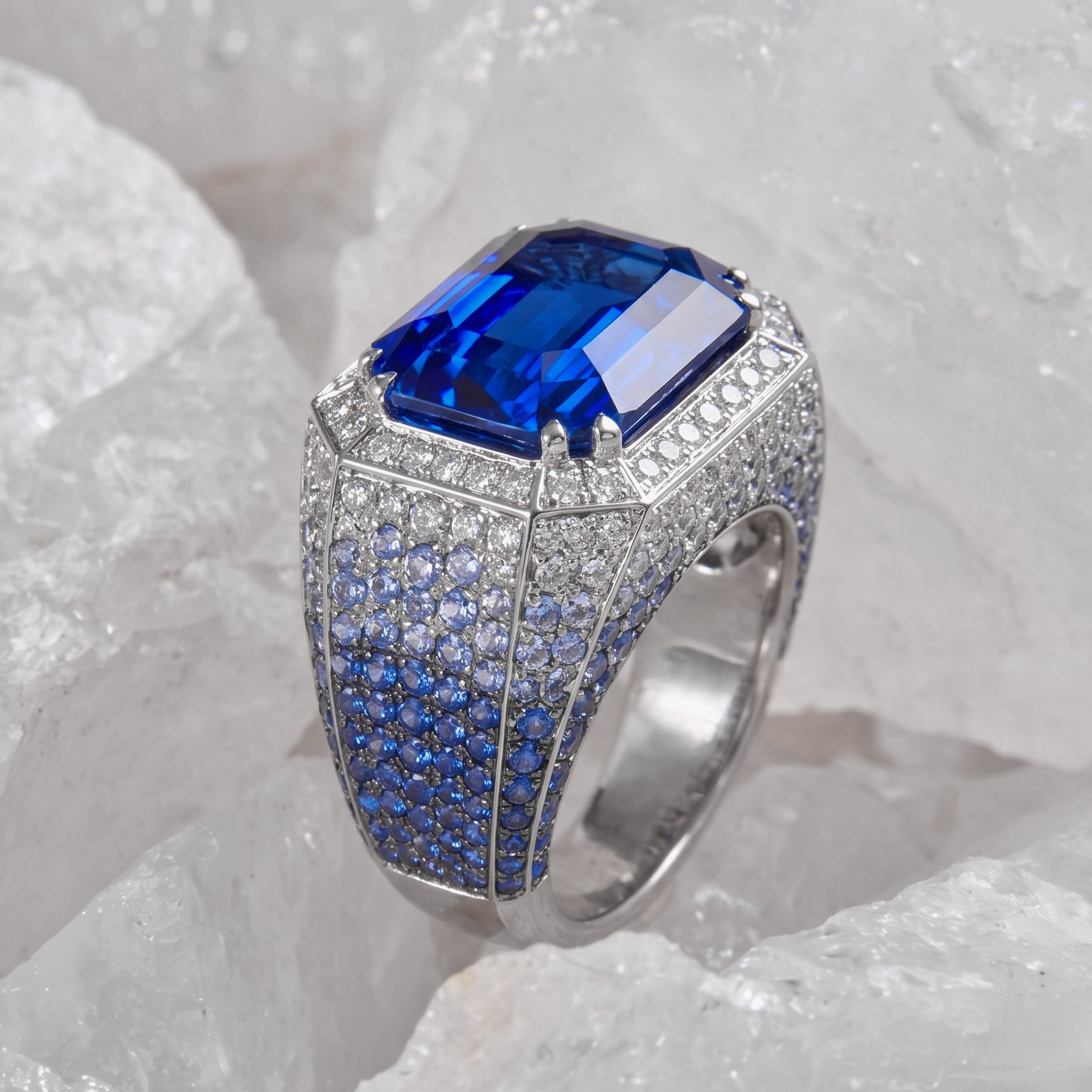 Large blue rectangular-cut gemstone ring with pavé diamonds along the band in a silver setting against a pale background.