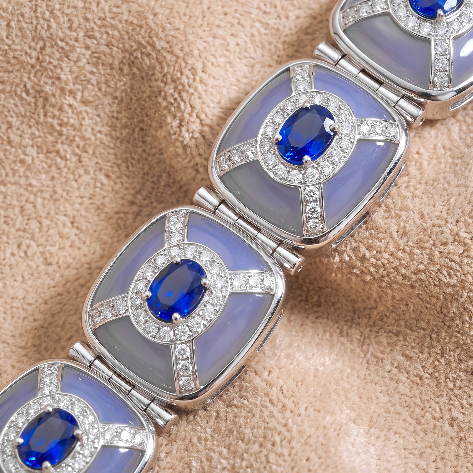 Silver bracelet links with oval blue gems surrounded by diamonds on a beige fabric background (close-up).