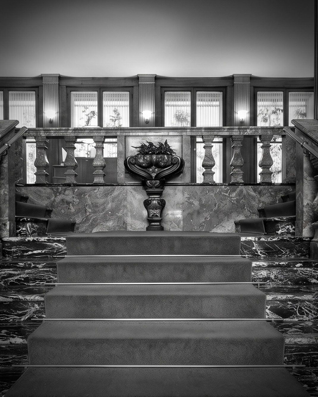 Marble staircase with an ornate balustrade and a decorative urn with plants on the landing, in a formal lobby (black and white).