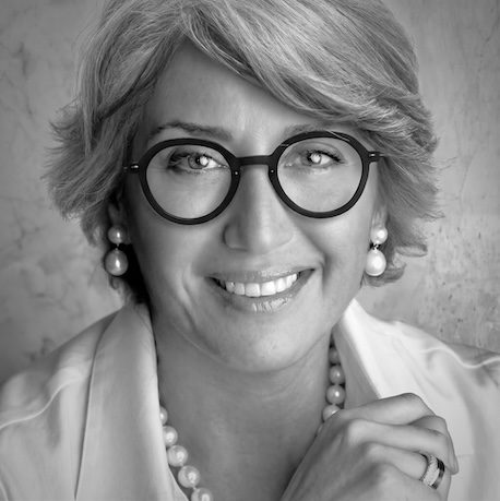 Smiling woman with short gray hair and round black glasses, wearing pearl earrings and necklace, hand resting near chin.
